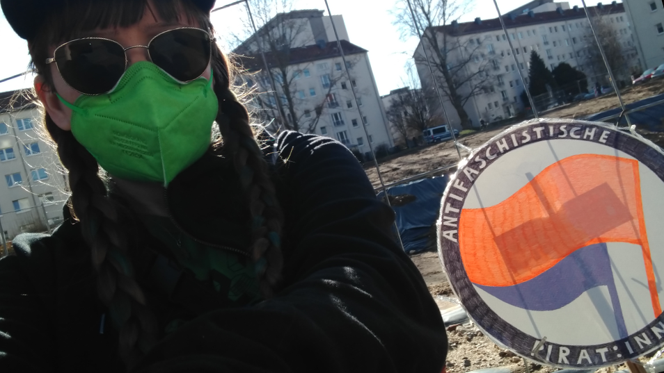 Ich mit Sonnenbrille, Maske und Zöpfen an einen Zaun gelehnt, neben mir ein Schild auf dem Antifaschistische Pirat:innen steht Antifaschismus Stephanie Henkel