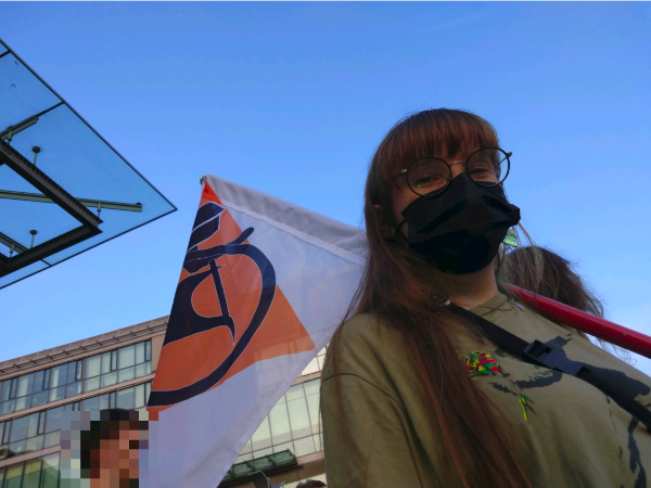 Ich mit einer schwarzen Maske und einer Fahne mit dem Logo der Hochschulpirat*innen darauf von schräg unten Demo Piraten
