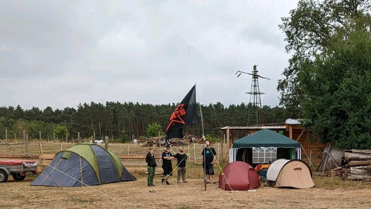 Menschen mit einer großen Fahne zwischen Zelten auf einer ausgetrockneten Wiese. Fedicamp