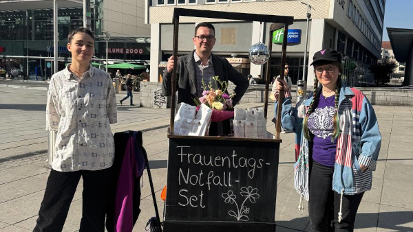 Drei Personen neben einem voll beladenen Handwagen in einer Einkaufsmeile. Fem_Kampftag_2025