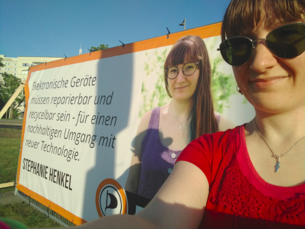 Ich vor einem Großplakat von mir auf dem steht: Elektronische Geräte müssen reparierbar und recycelbar sein - für einen nachhaltigen Umgang mit neuer Technologie. Stephanie Henkel Großplakat Stephanie Henkel Piraten