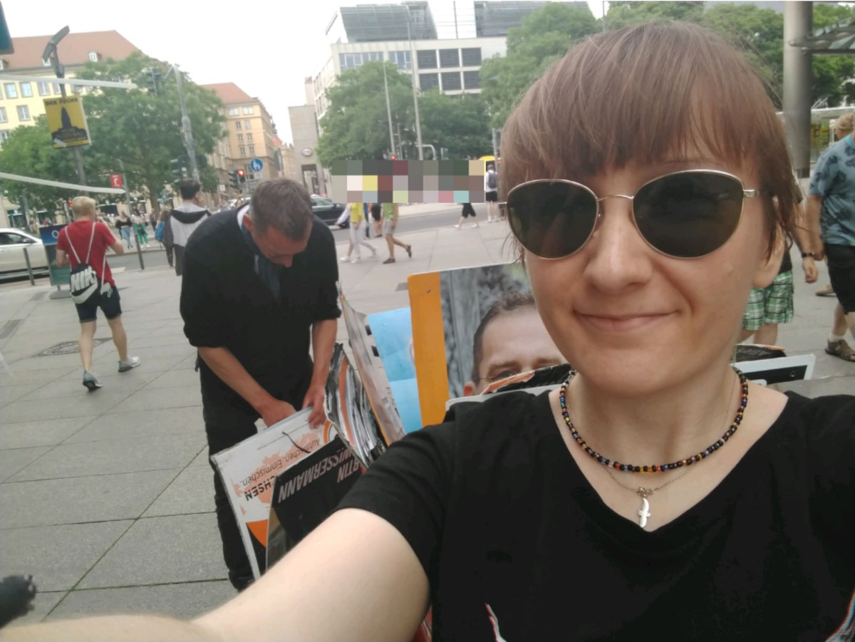 Ich mache mit Sonnenbrille auf der Nase ein Selfie auf einer belebten Einkaufsstraße im Hintergrund. Im Mittelgrund msw, wie er aus einem Anhänger heraus Plakate sortiert. MSW mit Ückück im Wahlkampf OBW22
