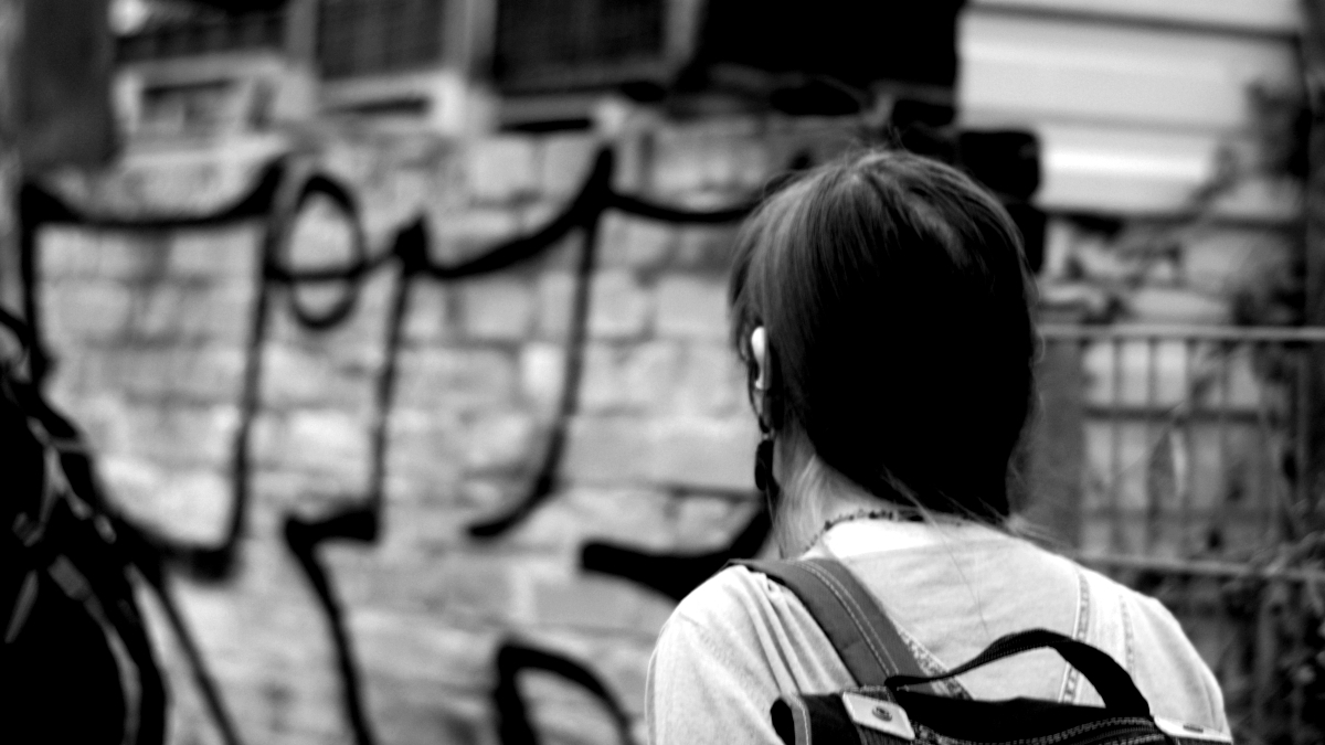Eine Fotografie in Grautönen von einer Person schräg von hinten, wie sie nach links in das Bild hinein schaut. Im Hintergrund bemalte Wände. Wenn%20nicht%20wir,%20wer%20dann